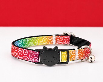 Könnte beinhalten: Ein Haustierhalsband mit einem lebendigen Regenbogen-Farbverlauf und weißen Wirbelmustern. Es verfügt über eine schwarze Kunststoffschließe in Form eines Katzengesichts, einen silberfarbenen Metall-D-Ring und eine kleine silberne Glocke an der Seite.