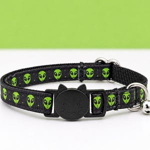 May include: A black nylon pet collar featuring a repeating pattern of small green alien heads. The collar includes a black plastic buckle shaped like a cat face with whiskers and ears, along with a silver metal D-ring and a small attached bell.