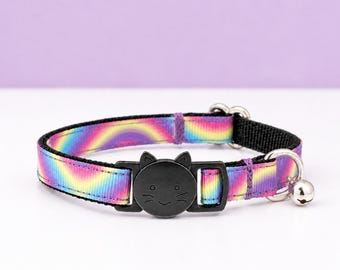 Könnte beinhalten: Ein Haustierhalsband mit einem leuchtend regenbogenfarbenen Wellenmuster auf einem Nylonband. Das Halsband verfügt über eine schwarze Kunststoff-Sicherheitssteckschnalle in Form eines Katzengesichts mit eingravierten Schnurrhaaren und eine kleine Metallglocke.