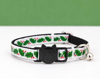 May include: A white pet collar featuring a repeating pattern of green holly leaves and red berries. The collar includes a black plastic buckle shaped like a cat face with whiskers and ears, along with a small silver metal bell attached to a ring.