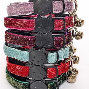 May include: A stack of six pet collars in various glittery colors including lavender, magenta, dark purple, light blue, red, and dark green. Each collar features a black plastic buckle shaped like a cat face and a small silver metal bell attached.