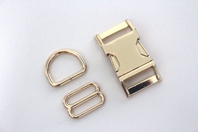 May include: A collection of three gold-coloured metal hardware components. The set includes a D-ring, a slide adjuster, and a side-release buckle. These findings are suitable for bag making, pet collars, or other craft applications.