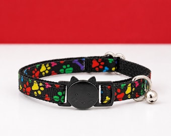Könnte beinhalten: Ein schwarzes Haustierhalsband mit einem bunten Muster aus roten, gelben, grünen und blauen Pfotenabdrücken. Das Halsband verfügt über eine schwarze Kunststoffschnalle in Form eines Katzengesichts mit Ohren und eine kleine silberne Metallglocke.