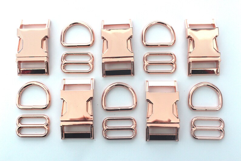 May include: Rose gold metal hardware set including buckles, D-rings, and tri-glide slides. The set is arranged on a white surface. The hardware is likely for use in crafting or pet accessories.