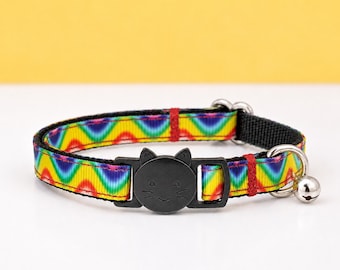 Könnte beinhalten: Ein Haustierhalsband mit einem leuchtenden Regenbogen-Zickzackmuster auf einem gewebten Band. Das Halsband verfügt über eine schwarze Kunststoffschnalle in Form eines Katzengesichts mit eingravierten Schnurrhaaren und Ohren sowie eine kleine silberne Metallglocke.