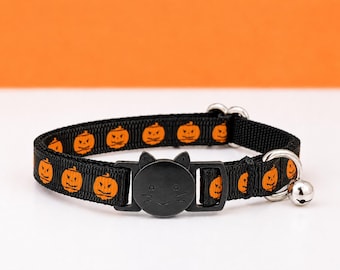 May include: A black nylon pet collar featuring a repeating pattern of orange jack-o'-lanterns. The collar includes a black plastic breakaway buckle shaped like a cat head with whiskers and a small silver-toned metal bell attached to a D-ring.
