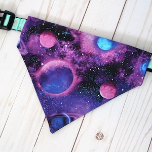 May include: A triangular dog bandana featuring a vibrant galaxy print with purple, pink, and blue planets, stars, and nebulae. The bandana has a black plastic buckle and a turquoise strap with a gold pattern.