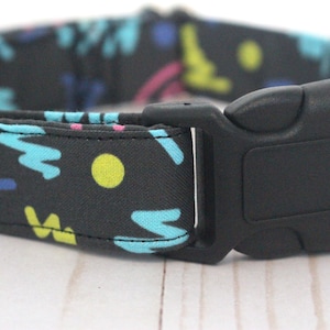 May include: A black dog collar with a colourful geometric pattern, featuring blue, yellow, pink, and green shapes. The collar has a black plastic buckle.