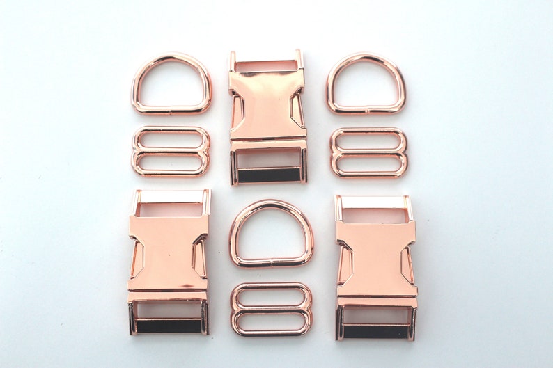 May include: Rose gold metal hardware set, including D-rings, slide adjusters, and side-release buckles. The hardware is arranged on a white surface, showcasing the shiny, metallic finish. Ideal for crafting pet collars, bags, or other accessories.