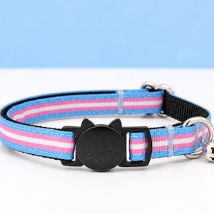 May include: A pet collar featuring a striped pattern in light blue, white, and pink. The collar includes a black plastic breakaway buckle shaped like a cat face with etched whiskers and eyes, along with a small silver-toned metal bell attached.