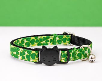 May include: A pet collar featuring a green and white shamrock pattern on a fabric strap with a black plastic buckle shaped like a cat face. The collar includes a small silver-toned metal bell attached to a metal D-ring and a black nylon backing.