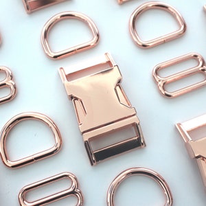May include: Rose gold metal hardware assortment, including D-rings, buckles, and sliders. The hardware pieces are arranged on a white surface, showcasing their shiny, metallic finish. Ideal for crafting and accessories.