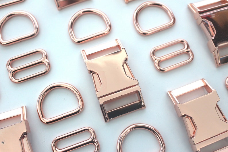 May include: Rose gold metal hardware assortment, including D-rings, buckles, and sliders. The hardware pieces are arranged on a white surface, showcasing their shiny, metallic finish. Ideal for crafting and accessories.