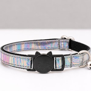 Könnte beinhalten: Schillernder Katzenhalsband mit schwarzer katzenförmiger Schnalle und kleiner silberner Glocke. Das Halsband hat einen reflektierenden, regenbogenartigen Glanz. Die Schnalle zeigt ein lächelndes Katzengesicht. Ein stilvolles und sicheres Accessoire.