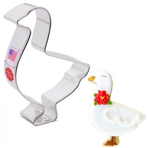 May include: A stainless steel goose-shaped cookie cutter next to a decorated goose cookie. The cookie is white with a red flower collar and a yellow beak. The cookie cutter has a "Made in USA" label.