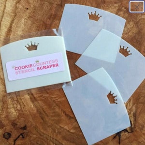 May include: A set of cookie stencils and a scraper. The scraper is off-white with a crown cutout and a label that reads "Cookie Countess Stencil Scraper". The stencils are clear with a crown cutout. The items are on a wooden surface.