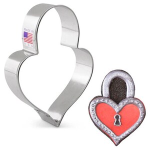 May include: A heart-shaped cookie cutter made of shiny, silver-colored metal. The cutter has a label that reads "MADE IN USA". Next to it is a heart-shaped cookie decorated to resemble a lock, with a red center and silver trim.