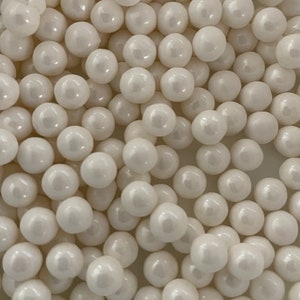 May include: A close-up image of a large quantity of white pearl-like candy sprinkles. The sprinkles are round and have a slight sheen.