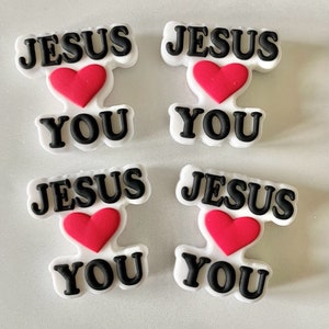 May include: Four white and black charms with the words "JESUS YOU" and a red heart in the center. The charms are in a block letter style and are likely shoe charms or accessories.