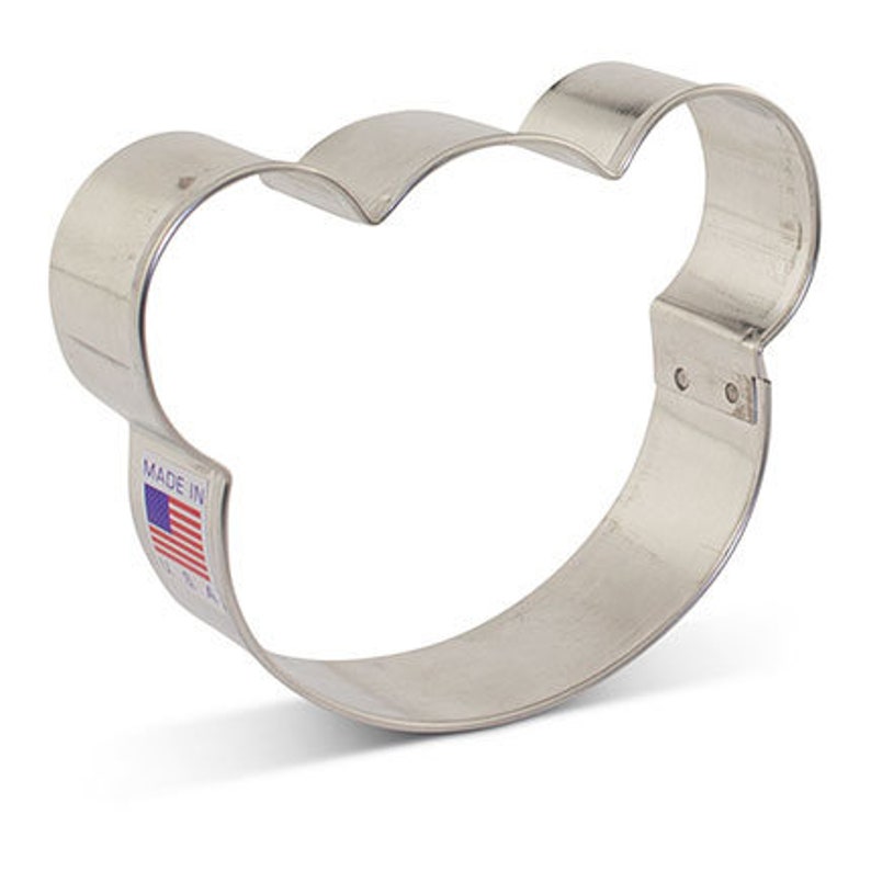 May include: A silver-colored metal cookie cutter in the shape of a bear's head. The cutter has a rounded shape with three curved sections at the top and two rounded sections on the sides. The words "MADE IN" and a small American flag are visible.