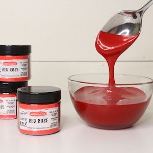 May include: A close-up shot shows jars of red food coloring labeled "Red Rose" next to a glass bowl filled with the same vibrant color. A spoon is used to pour the liquid into the bowl. The jars have black lids and red labels.