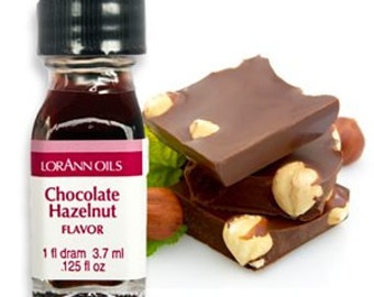 May include: A small glass bottle of chocolate hazelnut flavoring. The bottle is labeled "Lorann Oils Chocolate Hazelnut Flavor 1 fl dram 3.7 ml .125 fl oz". The bottle is sitting on a white surface with a pile of chocolate squares and hazelnuts.
