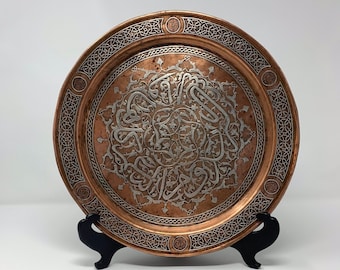 May include: A round copper tray with intricate silver filigree designs. The tray has a raised rim with a decorative border. The center of the tray features a circular design with Arabic calligraphy.