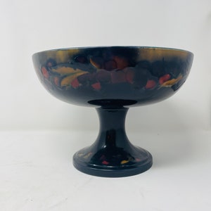 Peut inclure: Un bol sur piédestal foncé avec un large bord et une base évasée courte. Le bol est noir avec une finition brillante et présente un motif décoratif de feuilles et de baies rouges et dorées autour du bord.