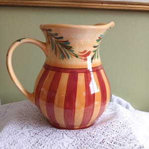 May include: A ceramic pitcher featuring a glossy finish with a cream-colored upper section decorated with painted green and red leaf motifs. The lower body displays vertical stripes in alternating shades of deep red and warm orange-yellow.