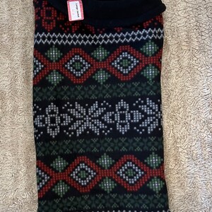 May include: Black fleece dog sweater with a red, green, and white geometric pattern. The sweater has a black turtleneck collar and a tag that reads "Large Jacket 16-18"