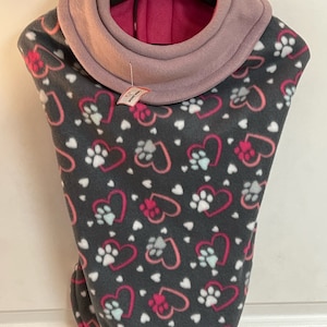 May include: A gray fleece dog sweater with a pink and purple fleece collar. The sweater has a pattern of pink and white hearts with paw prints inside the hearts. The sweater is on a hanger.