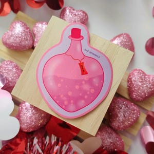 May include: A pink potion bottle sticker with a red stopper and a small tag. The sticker is on a wooden surface, surrounded by pink glitter hearts and red decorations. The text "KTZ ORIGINALZ" is visible.