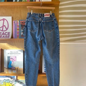 Puede incluir: Vaqueros de mezclilla azul claro colgados de una percha de madera. Los vaqueros tienen un diseño clásico de cinco bolsillos con un parche "BONGO" en la cintura trasera. Los vaqueros se exhiben sobre un fondo de estantería y pared a rayas.