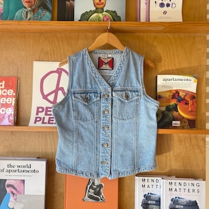 May include: A light blue denim vest with a V-neck and button-up front hangs on a wooden hanger. The vest has two chest pockets and a small label. The background features a wooden shelf with magazines and books.