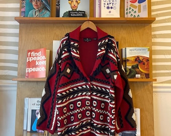 May include: A red, white, and black patterned cardigan hangs on a wooden rack with several magazines. The cardigan features a geometric design and a button-up front. The magazines on the shelves have various covers and titles.