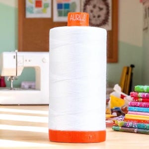 May include: A large spool of white sewing thread with an orange base and top. The brand name "AURI" is visible on the top. A sewing machine and colorful fabric scraps are in the background. Ideal for sewing projects.