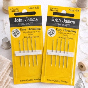 May include: Two packages of John James Easy Threading needles, size 4/8, each containing six needles. The yellow packaging includes text in English, French, and German. Other sewing notions, including spools of thread and buttons, are visible.
