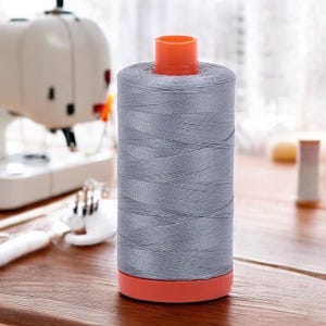 May include: A spool of gray thread with an orange top and base. The thread is tightly wound, and the spool is cylindrical. The background includes a sewing machine and other sewing supplies, suggesting a crafting or sewing theme.