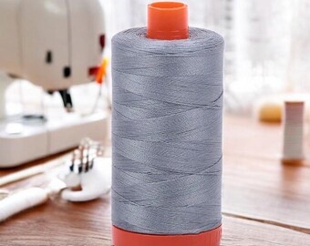 May include: A spool of gray thread with an orange top and base. The thread is tightly wound, and the spool is cylindrical. The background includes a sewing machine and other sewing supplies, suggesting a crafting or sewing theme.