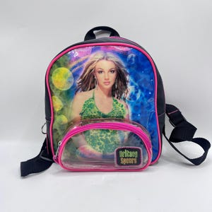 May include: A black mini backpack featuring a vibrant graphic print of Britney Spears wearing a green patterned top. The bag has a clear front pocket with a pink zipper trim and a rectangular patch that reads Britney Spears in colorful letters.