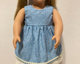 May include: A blue and white patterned dress with a white lace trim. The dress is sleeveless and has a gathered skirt. The doll is wearing the dress.