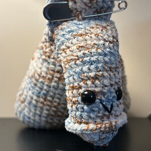 May include: A crocheted plush toy in the shape of a worm with blue, white, and brown yarn. The toy has black eyes and a small black mouth. A safety pin is attached to the top of the toy.