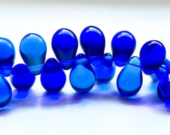 May include: A strand of bright blue teardrop-shaped glass beads. The beads are strung together on a clear thread and are arranged in a curved line.