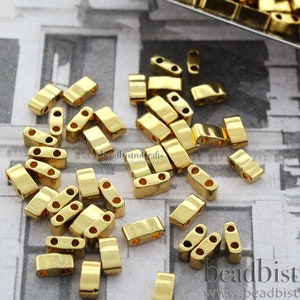 May include: Gold-colored metal beads with two holes each. The beads are arranged in a pile on a white and gray patterned surface. The beads are approximately 5mm in length.