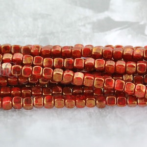 May include: A strand of small, square, red and gold metallic beads. The beads are arranged in a row and are close together. The beads are shiny and have a metallic finish.