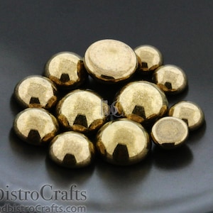 May include: A collection of gold-colored round cabochons, also known as flat-back gems, on a black surface. These cabochons are ideal for jewelry making, crafting, and embellishing projects.