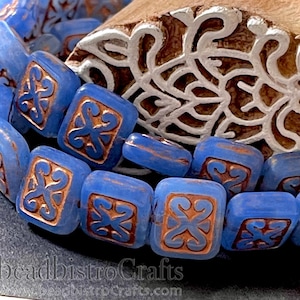 May include: Blue glass square beads with a brown swirl design. The beads are stacked on top of each other and are in focus. The background is out of focus and shows a wooden block with a carved design.