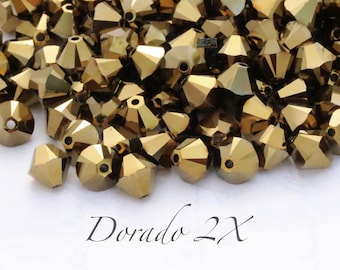 May include: A pile of gold-colored glass beads with a faceted, conical shape. The beads are arranged in a random pattern and are all the same size. The text "Dorado 2X" is written in cursive on the white surface below the beads.