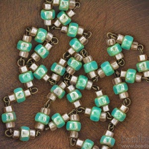 May include: A turquoise and silver beaded necklace with a square shape. The beads are strung on a silver wire and have a metallic sheen.