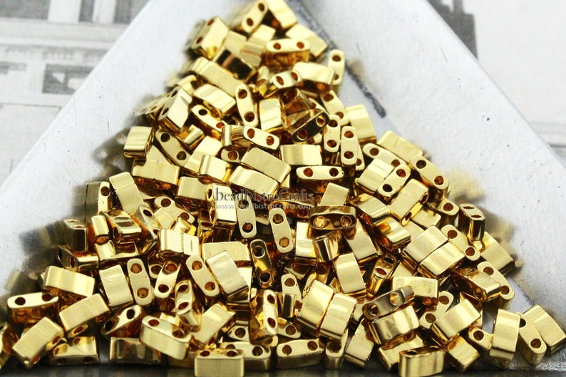 May include: A pile of gold-colored metal beads with two holes in each bead. The beads are rectangular in shape and have a shiny finish.